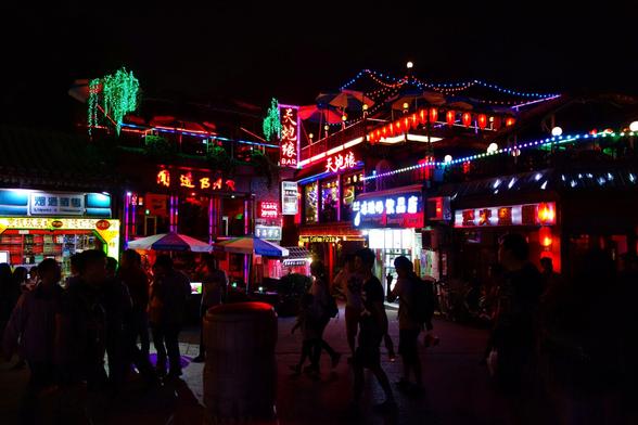 A vibrant night-time street scene with multiple buildings decorated with bright, colourful neon lights and traditional red lanterns. People are walking along a busy street, some in groups and others individually, with a large, decorative wooden barrel in the foreground. The buildings have signs with Chinese characters that are lit up in red and blue neon. The atmosphere is lively and bustling, with a clear night sky visible in the background.
