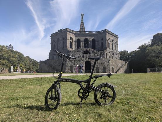 Das Fahrrad "Birdy" von Riese und Müller steht auf einer Wiese und im Hintergrund ist der Herkules (Kassel) von hinten zu sehen