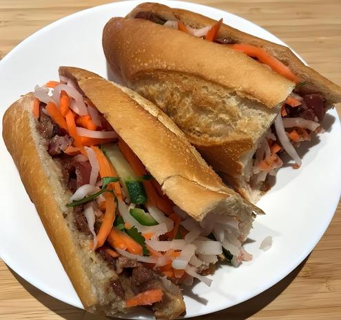 Homemade bánh mì sandwich, cut in half on a white plate, resting on wood dining table. The bánh mì has the classic crusty baguette. The sandwich in the foreground is cut in half, revealing the filling: thinly sliced garlic fried pork, pickled carrots, daikon radish & chopped jalapeño peppers. There are also slices of cucumber and cilantro.