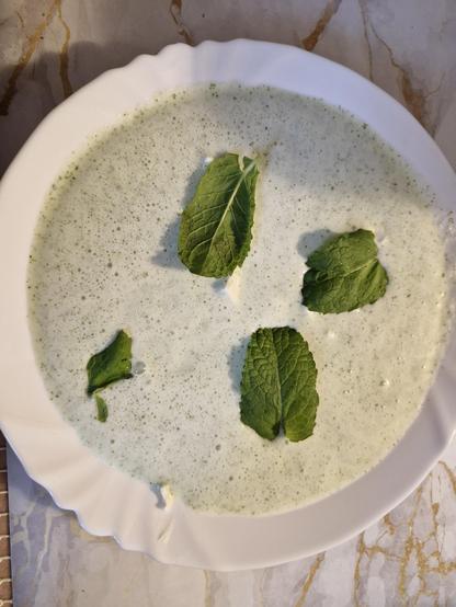 Na zdjęciu znajduje się biały, okrągły talerz ustawiony na stole pokrytym marmurowym blatem w jasnych odcieniach beżu i szarości. Na talerzu widoczna jest jasna, lekko zielonkawa, kremowa zupa lub napój, najprawdopodobniej chłodnik lub jogurt z dodatkiem ziół. Konsystencja potrawy jest płynna i gładka, z drobnymi zielonymi kropeczkami, sugerującymi zblendowane liście ziół. Na powierzchni zupy ułożone są cztery świeże zielone listki mięty, rozmieszczone w sposób umiarkowanie nieregularny: jeden bliżej środka, trzy pozostałe – bliżej brzegów talerza. Obrzeże talerza jest lekko falowane. Całość sprawia wrażenie orzeźwiającej i lekkiej potrawy, dekoracyjnie podanej.