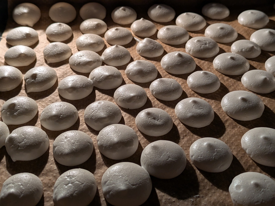 Auf dem Bild sehen wir viele kleine, weiße Baisers, die auf einem Backblech mit Backpapier liegen. Die gleichmäßige Form und die glänzende Oberfläche deuten darauf hin, dass sie frisch aufgespritzt und gerade gebacken oder getrocknet wurden.