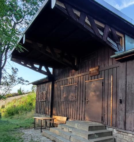 Ein Holzhaus mit 4 Stufiger Treppe vor der Eingangstür und einem großem Vordach darüber. Ein Tisch und eine Bank stehen vor dem Haus mit unter dem Vordach.
