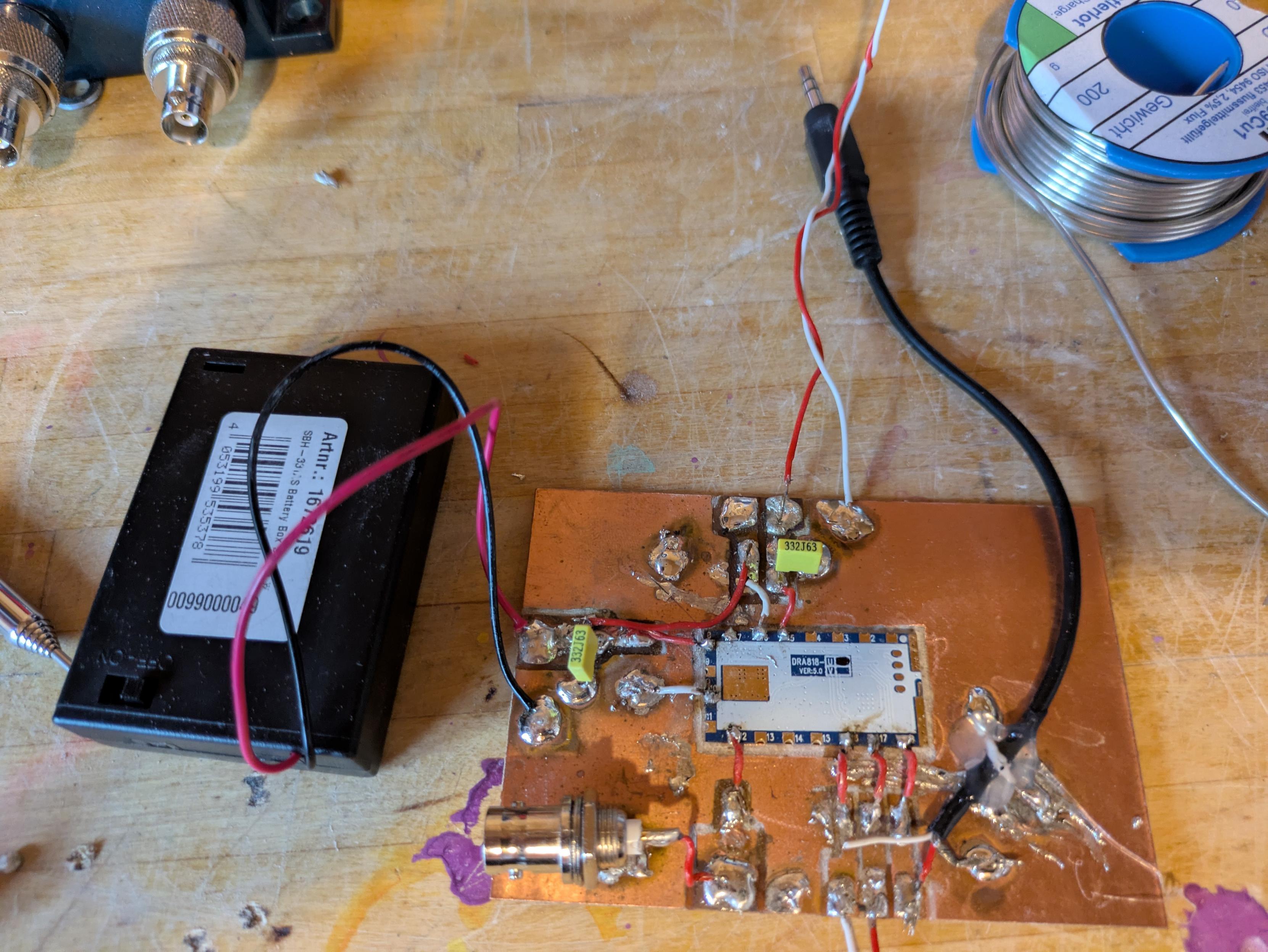 A hand carved copper.pcb with a vhf module in the middle and power, UART, Audio and.PTT cables  leading to carved out.islands. A BNC connector is soldered onto the pcb and a battery and audio cable as well