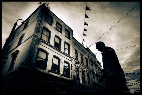 Dramatic low-angle black and white photograph of weathered buildings on North Main Street, Cork with silhouetted figure walking past, featuring decorative bunting strung between buildings and moody overcast sky.