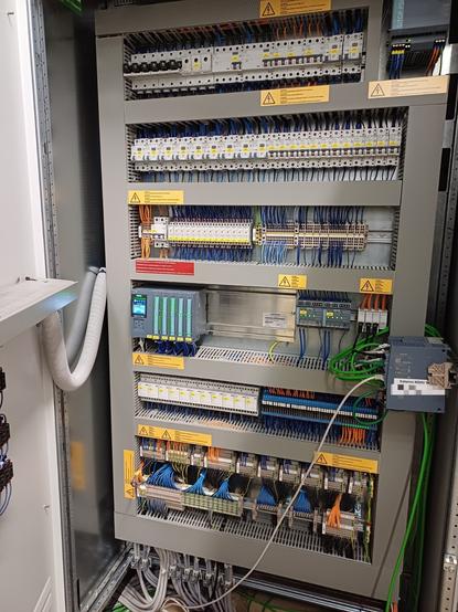An open electrical control cabinet with multiple rows of components. At the top are miniature circuit breakers (MCBs) and residual-current devices (RCDs). Below them are terminal blocks with organized blue and orange control wiring. On the left side is a Siemens SIMATIC S7 PLC with several I/O modules. On the right side are Siemens industrial Ethernet switches. A Siemens Sitop power supply is also installed. Green Ethernet and fieldbus cables are connected to the switches, while various control and power cables exit the cabinet at the bottom through conduits. Several yellow warning labels are attached to indicate electrical hazards.