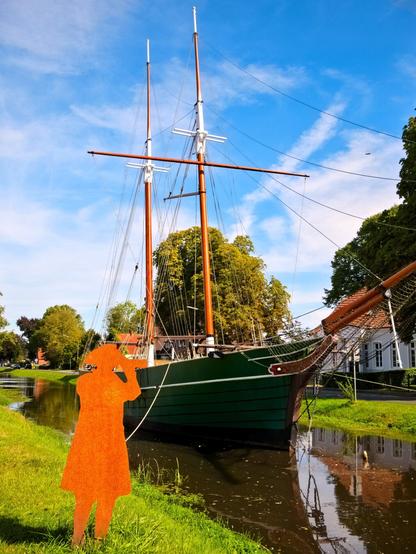 A sailing ship with two masts floats on a canal; the hull of the ship is green. In the foreground stands a rusty figure wearing a hat and coat, holding binoculars and looking at the ship. The ship is the ‘Catharina von Papenburg’. The sky is blue with a few clouds. The sun is shining.
