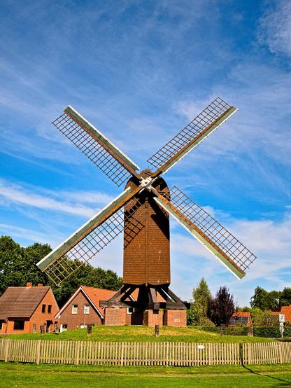 A windmill on a hill. In the background, red brick houses. The mill has a narrow body and four sails.