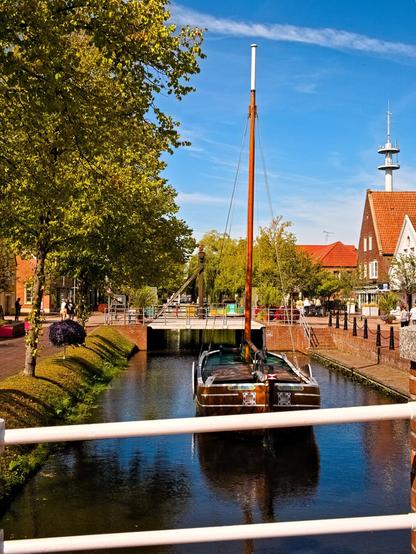 A canal in the sunlight. A ship with a mast lies there. In the background, a bridge and small-town life.
