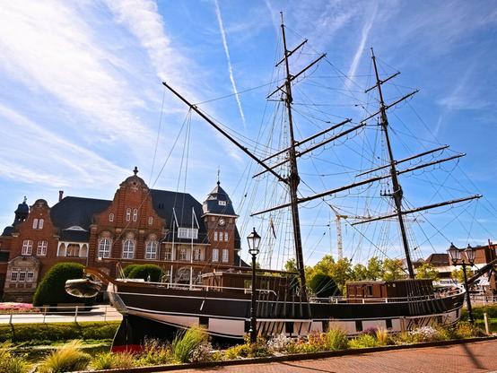 Another canal, another ship. This one has two masts and a green and white hull. A pretty building in the background. Sunshine.
