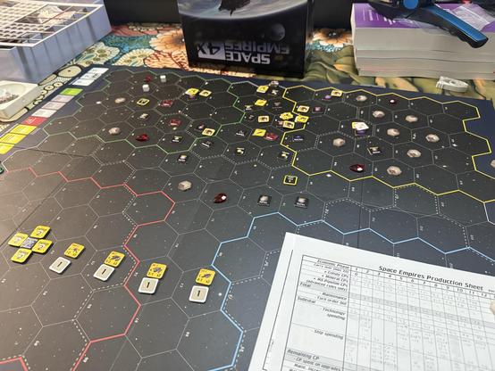 A tabletop game setup featuring a hexagonal game board with various colored tokens and resources. Nearby, there is a production sheet detailing game mechanics. A game box and a stack of books are visible in the background. The board depicts a space exploration theme.