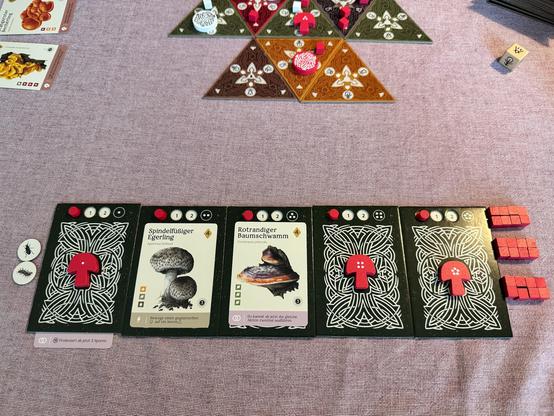 Das Bild zeigt ein Brettspiel mit mehreren Karten und Spielteilen auf einem Tisch. Im Vordergrund liegen fünf Karten mit verschiedenen Pilzarten ausgerichtet, darunter "Spindelfüßeriger Egerling" und "Rotrandiger Baumschwamm". Jede Karte hat ein rotes Symbol und Punkte, die wahrscheinlich die Spielwerte darstellen. Neben den Karten liegen rote Spielsteine und kleine weiße Münzen. Im Hintergrund sind mehrere dreieckige Spielplatten mit verschiedenen Farben und Symbolen zu sehen, auf denen rote und weiße Spielsteine platziert sind. Ein Würfel mit einem Symbol ist ebenfalls sichtbar. Die Karten und Spielteile deuten auf ein strategisches Brettspiel hin, das möglicherweise mit Pilzen und Naturthemen zu tun hat.