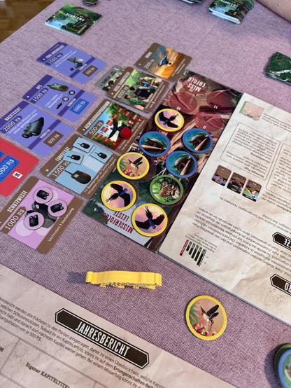 Das Bild zeigt eine Tischszenario-Spiel-Setup auf einem lilafarbenen Tisch. Im Vordergrund liegen verschiedene Spielkarten und Token, die mit verschiedenen Werten und Illustrationen versehen sind. Die Karten sind in verschiedenen Farben wie Blau, Rot, Gelb und Braun und zeigen verschiedene Objekte und Tiere, wie zum Beispiel ein Vogel und ein Elefant. Einige Karten haben Preise in "RS" angegeben, wie z.B. "2000 RS" und "1500 RS". Einige Karten zeigen auch Text in Deutsch, wie "Mittelpunkt" und "Schutzweste". Im Hintergrund ist ein Spielbrett mit einem Titel "Jahresbericht" zu sehen, der auf Deutsch ist. Es gibt auch mehrere Spielzockel mit Tierillustrationen, wie zum Beispiel ein Vogel und ein Schmetterling. Die gesamte Szene wirkt wie ein Spiel, das mit Themen aus der Natur und Forschung zu tun hat.