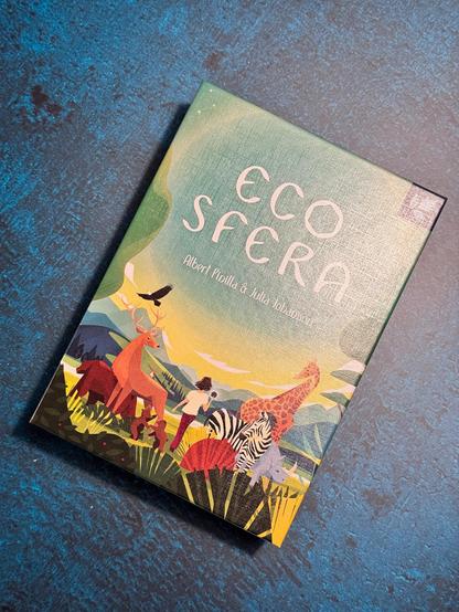 Das Bild zeigt das Cover von "Eco Sfera" von Albert Pintilla und Julia Johansson. Das Cover ist in leuchtenden Farben gestaltet und zeigt eine idyllische Landschaft mit verschiedenen Tieren wie einem Reh, einem Giraffen, einem Zebra und einem Nashorn. Im Vordergrund steht eine Person, die eine Blume hält, und ein Adler fliegt in der Luft. Der Hintergrund zeigt sanfte Hügel und ein farbenfrohes Himmelblau. Der Titel "Eco Sfera" ist in großen, weißen Buchstaben auf dem Cover zu sehen, und die Namen der Autoren stehen darunter. Das Buch liegt auf einem blauen, texturierten Untergrund.
―
