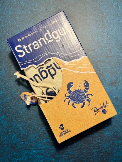Das Bild zeigt eine Verpackung für ein Spiel namens "Strandgut" von Bryan Burgoyne und Fanny Saulnier. Die Verpackung ist in zwei Farben gehalten: oben ist ein blauer Bereich mit dem Titel "Strandgut" in großen, weißen Buchstaben, und unten ein brauner Bereich mit einem blauen Karabinerfisch und dem Namen "Randolph" in einer geschwungenen Schrift. Der Titel "Strandgut" ist auch auf einem beige-braunen Stoffbeutel mit einem Zugband zu sehen, der teilweise aus der Verpackung hervorragt. Der Stoffbeutel trägt die gleiche Schrift wie auf der Verpackung. Die Verpackung ist auf einem blaugrünen, texturierten Untergrund platziert. Im unteren linken Bereich der Verpackung steht "Edition Spielweise," was auf eine spezielle Ausgabe hinweist.