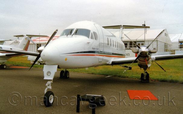 Forward view of a twin propellor-engined airliner parked on a taxiway facing slightly to the left of the camera.
The plane is mostly white, with a black and orange stripe running along the body, and the words "Hawker Pacific" just below the cockpit side windows.
There is a red mat on the floor, just in front of the plane's left engine, and a small black step beside the nose-wheel.
Green grass in the background leads up to a series of white pre-fabricated buildings, under grey skies.