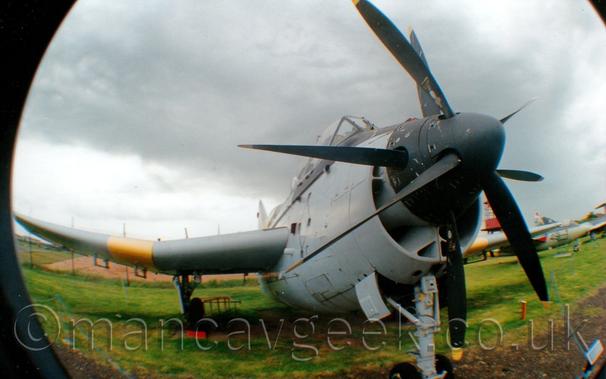Front view of a twin propellor-engined, short and stubby military aircraft on display parked on concrete pads in the middle of a large grassed area, taken with a semi-fish-eye converter on a wide-angled lens, a combination that distorts straight lines while allowing a greater field of view at a closer range..
The 2 engines drive a set of contra-rotating propellors on the nose, each driving a different propellor on the same shaft, which explains the 2 sets of propellor blades.
The plane is largely a pale grey-blue, with black around the cockpit canopy and around the propellors in the nose.
There are yellow bands around the wings, just outboard of the main landing gear.
Green grass fills the bottom of the frame, with more military planes in the background on the right.
Dramatic grey sky fills the top of the frame.