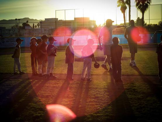 The image depicts a group of children and two adults on a baseball field during sunset. The children, wearing baseball caps and some with gloves, are standing in a semi-circle, facing the adults. The adults are positioned on the right side of the image, with one gesturing towards the children. The field is surrounded by a fence, and palm trees are visible in the background, indicating a warm climate. The sun is low in the sky, creating a warm glow and lens flare effects, casting long shadows on the ground. The scene suggests a moment of instruction or discussion, possibly after a baseball game or practice.

Provided by @altbot, generated privately and locally using Ovis2-8B