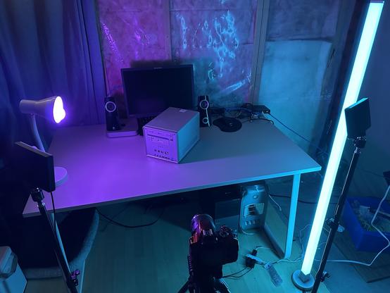 A moody, retro-tech filming setup lit with nostalgic, dramatic cyan and magenta lighting. A beige IKEA desk is positioned against an unfinished wall with exposed insulation. On the desk sits a silver Shuttle XPC cube-style PC, illuminated with pink and blue light, in front of a Lenovo ThinkVision monitor. A pair of Logitech speakers, a wired keyboard, a black mouse, a network switch, and a Mac Mini are also on the desk. To the left is a purple desk lamp, and on both sides in front of the desk are two LED lights mounted on tripods, but only the tall cyan light stick and the purple desk lamp are turned on. A camera is set up on a tripod in front of the desk, aimed at the Shuttle PC. Underneath the desk sit a silver Power Mac G4 MDD and a Dell Optiplex tower.