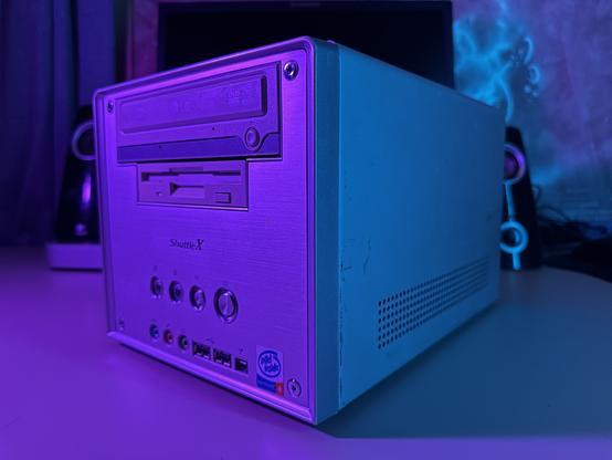 A close-up of the silver Shuttle XPC cube computer, dramatically lit with magenta and cyan lighting. The front panel features a DVD-ROM drive, a floppy drive, and front I/O ports including USB, audio jacks, and power/reset buttons. A classic blue Intel Pentium 4 “Intel Inside” sticker is visible in the lower right corner. The metal case has some wear and scratches, showing its age. The background is lightly out of focus and blurred, but still shows the black Lenovo monitor, desktop speakers, and the wall’s insulation behind the desk.