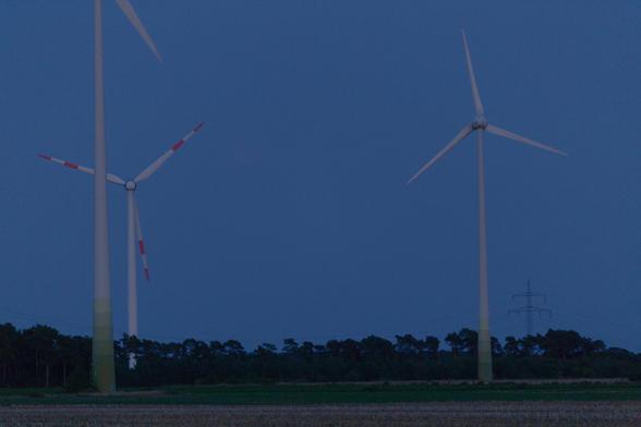 Foto des Dämmerungshimmel mit drei Windrädern und Wald am Horizont. Der verfinsterte Mond ist kaum zu erkennen.