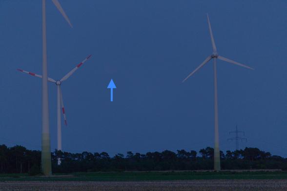 Photo of the twilight sky with three wind turbines and forest on the horizon. The eclipsed moon is barely visible and is therefore marked with a blue arrow in the image.