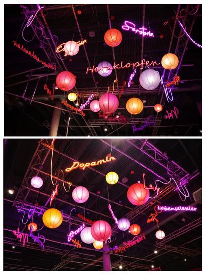 A vertical diptych showing a close-up of a ceiling installation from two slightly different angles. The installation features a collection of pink, orange, yellow, and purple paper lanterns hanging at varying heights. Interspersed among the lanterns are several neon signs spelling out German words in a fluid, script-like font. In the top image, words like 'Herzklopfen' (heartbeat) and 'Endorphin' are visible. The bottom image shows words such as 'Dopamin' (dopamine), 'Stress', and 'Lebenselixier' (elixir of life). The entire setup is suspended from a grid-like metal structure, and the background is dark, drawing focus to the colourful, illuminated display.