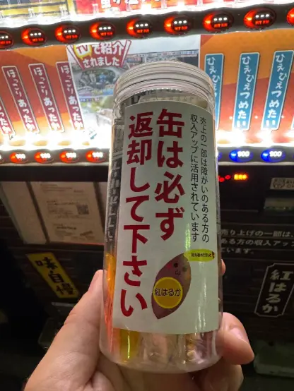 A hand holding a clear cylindrical container with Japanese writing and an illustration of a sweet potato on it. The background shows a vending machine offering more of these delicious tubes.