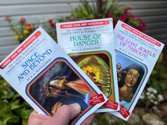 A hand holds three "Choose Your Own Adventure" books by R.A. Montgomery: "Space and Beyond," "House of Danger," and "The Lost Jewels of Nabooti." The background features vibrant flowers and greenery.