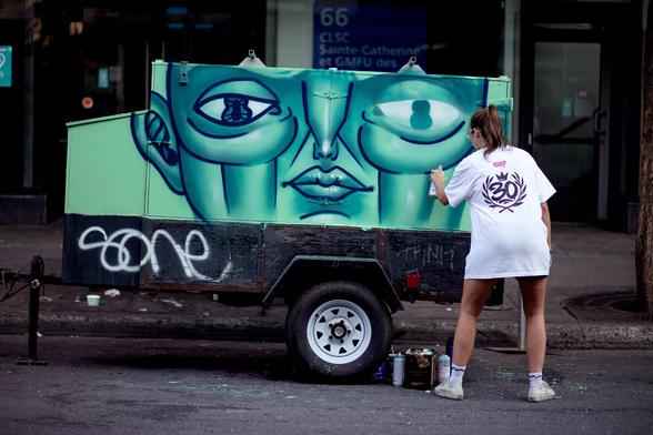 A young woman painting on an urban infrastructure in the street of Montreal. She is painting a green face. She is wearing a big white t-shirt of the UnderPressure festival that gives a nice contrast in the image.