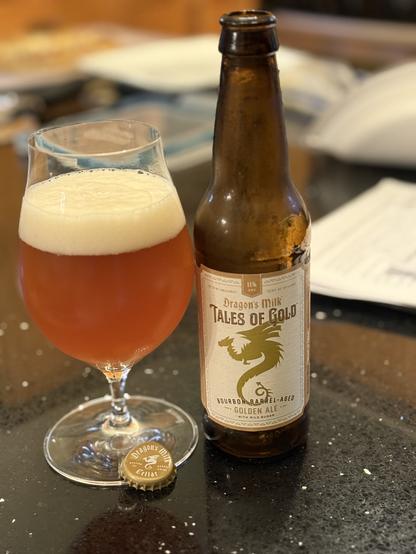 A snifter glass full of golden ale with a white head is next to a brown bottle with a white label white gold letting and a dragon in silhouette with its wings unfurled. Various clutter on the countertop is out of focus in the background.