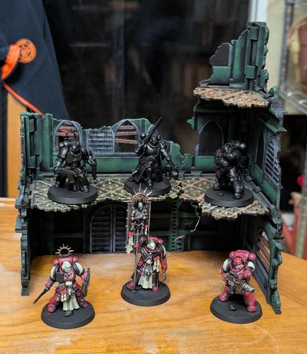 Painted Warhammer 40k minis on a piece of scenery. Three Space Marines painted as Blood Ravens and three more primed black in front of a green ruin.