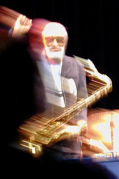 A blurred image of a saxophonist pumping his right fist up next to his head, wearing sunglasses and a suit. He has white hair and beard. The background is a dark performance stage, with faint outlines of drums visible. The motion creates a dynamic, energetic effect.