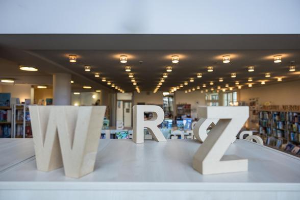 Blick über einen Regal in die Bibliothek. Darauf große Buchstaben aus Pappe.