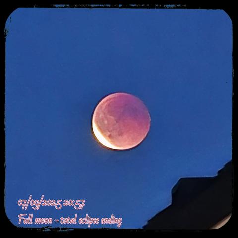 AI Alt Text:
A striking photograph of the moon during the final moments of a total lunar eclipse, taken on September 7th, 2025, at 20:57. The sky is a deep twilight blue, providing a smooth backdrop for the celestial event. The moon dominates the center of the image, glowing with a reddish tint from the eclipse, while a bright golden-white crescent begins to emerge along its lower left edge as Earth’s shadow recedes. The contrast between the soft red glow of the shadowed moon and the brilliant sliver of illuminated surface creates a dramatic, otherworldly effect. In the lower right corner, part of a silhouetted rooftop is visible, grounding the scene with a touch of the earthly perspective. Decorative text is overlaid at the bottom of the image, reading: “07/09/2025 20:57 – Full moon – total eclipse ending.”