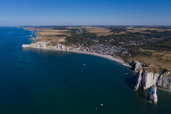 Aerial view of a coastal town with white cliffs and a sandy beach, surrounded by lush green fields and blue waters. Several boats are visible in the water.