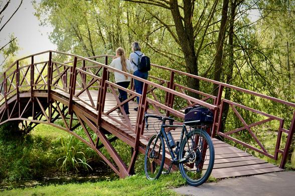 A wooden bridge spans a serene setting, with two people walking across it. Nearby, a bicycle is parked on the grass, surrounded by greenery and trees.