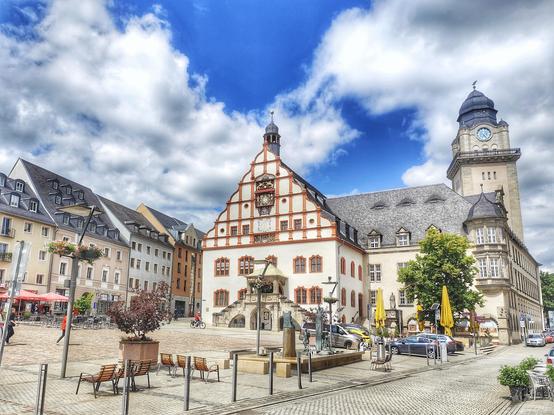 Historisches Rathaus in Plauen, dahinter ein Haus, das vom Rathausturm überragt wird. Vor dem Rathaus ein Platz mit Springbrunnen, links eine Häuserzeile. Der blaue Himmel bewölkt.