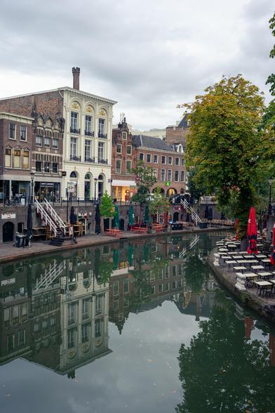 The image captures a tranquil canal scene in a European city on a cloudy day. On the left, multi-story buildings with varying facades—one white with decorative elements, another with dark brick and smaller windows—line the canal. A staircase leads down to the canal level, where a few people are seated beside a statue. To the right, a large tree stands next to an outdoor café area with neatly arranged tables and chairs. The calm canal reflects the buildings and foliage, creating a symmetric and serene atmosphere, despite the overcast sky.