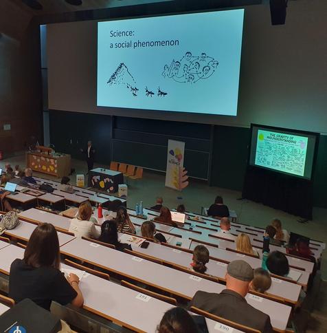 Photo Lecture Hall with ascending rows, from above, large screen "Science: a social phenomenon", people listening to speaker, small screen with graphic recording