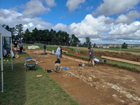 Scavo archeologico in corso con diversi archeologi al lavoro su un'area di terreno all'aperto, circondati da strumenti e materiali da costruzione, sotto un cielo parzialmente nuvoloso.