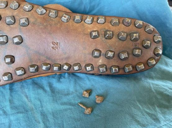 Immagine di una suola di scarpa in cuoio con chiodi di metallo, posizionata su un tessuto blu, con tre chiodi di ferro disposti accanto.