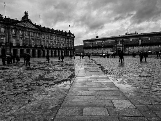 A wide-angle, black and white photograph of a large, wet cobblestone square. A straight path of flat stone slabs leads from the foreground into the distance. On the left, a large, ornate baroque building with many windows and statues on its roof dominates the view. On the right, a plainer, smaller stone building stands opposite. The square is scattered with people, some holding umbrellas, suggesting a recent rain shower. The wet cobblestones reflect the cloudy, dramatic sky. The overall scene is one of historical grandeur, captured on a dramatic, overcast day.