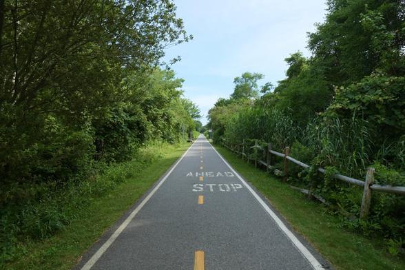 Color photo — a very straight, smoothly paved bike path leads through sunlit trees and low, dense greenery into the far distance. A yellow-painted dashed line separates the path into two lanes. Thick white lines mark the left and right edges of the path, with closely trimmed grass from the edges of the path to greenery on either side. An unpainted two-rail rustic wooden fence bounds the greenery on the right, leading to a distant red octagonal Stop sign — which marks a small crossing path. ‘STOP’ is painted below ‘AHEAD’ in white on the path just ahead. Minuscule figures — perhaps cyclists — are barely visible on the path in the far distance. The 14.3 mile path follows the former Providence and Bristol Railroad railbed, opened as a paved bike path in 1992,