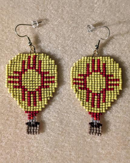 A pair of beaded earrings in the shape of hot air balloons; their design is a spin on the New Mexico flag (a red zia over a yellow background).