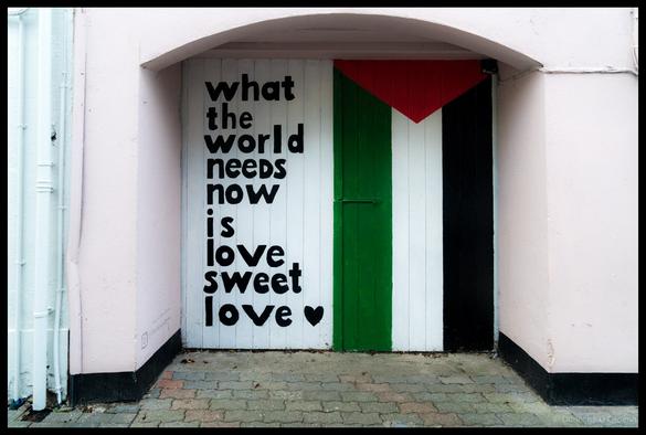 Arched white doorway in Kinsale, Co. Cork, featuring black text reading "what the world needs now is love sweet love" with a small heart symbol and a Palestinian flag painted in red, green, white and black stripes on wooden door panels.