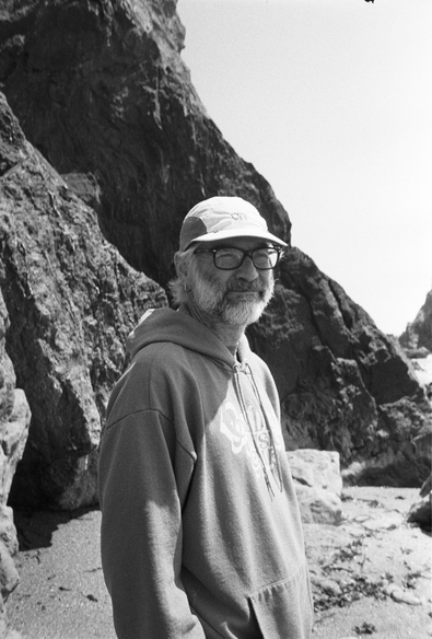 A black and white photo of a white-bearded man in a cap and hoodie at the beach.