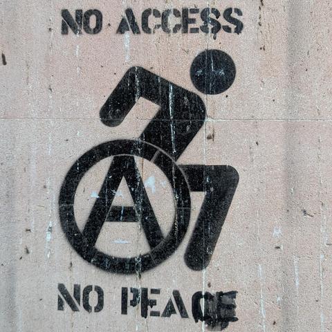 Un tag noir au pochoir sur un mur couleur brique "Pas d'accès pas de paix" en anglais, avec un logo fauteuil roulant actif, dont la roue est un A cerclé.