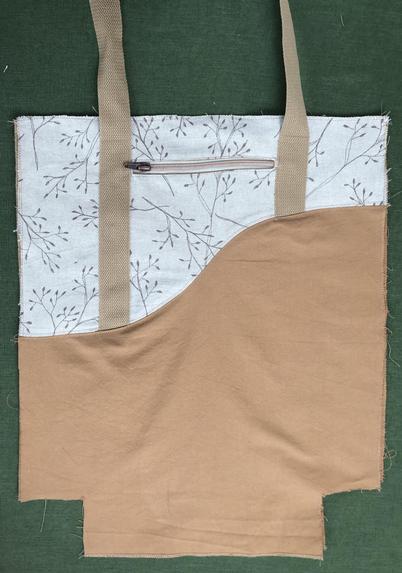 Halbe Einkaufstasche aus Stoff liegt auf grünem Tuch. Die Taschenhälfte ist durch eine bogenförmige Linie getrennt, in der oberen Hälfte ist sie beige und ocker, Gurtband ist sichtbar. Unterer Teil ist ocker Stoff, das Gurtband ist im unteren Teil nicht sichtbar.