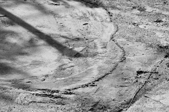 The shadow of a post rests on a sandy area covered in tracks and swirls, which creates an abstract composition.