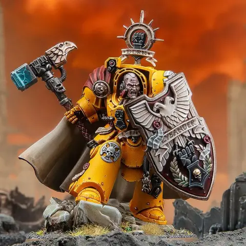 A dynamic miniature of Darnath Lysander in bright yellow Terminator armour, holding the relic storm shield Rampart in one hand and the Fist of Dorn power hammer in the other. His expression is fierce, and he wears a flowing cape, with intricate details on his armour and shield, including the name "LYSANDER" and various symbols. The background features a dramatic, fiery sky, enhancing the imposing presence of the character.